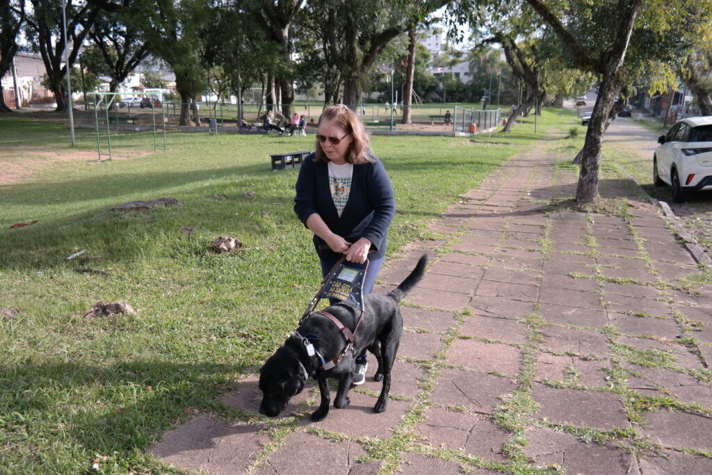 Uma mulher de cabelos claros e óculos escuros caminha por uma calçada de pedras acompanhada por um cão-guia preto, que segue à sua esquerda. Ela segura o arreio do animal enquanto percorre uma área arborizada. Ao redor, há gramado, árvores de grande porte e um parque ao ar livre.